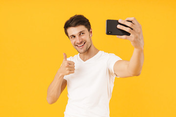 Happy young cheerful man in casual white t-shirt take a selfie by mobile phone isolated over yellow...