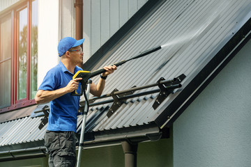 man standing on ladder and cleaning house metal roof with high pressure washer - obrazy, fototapety, plakaty