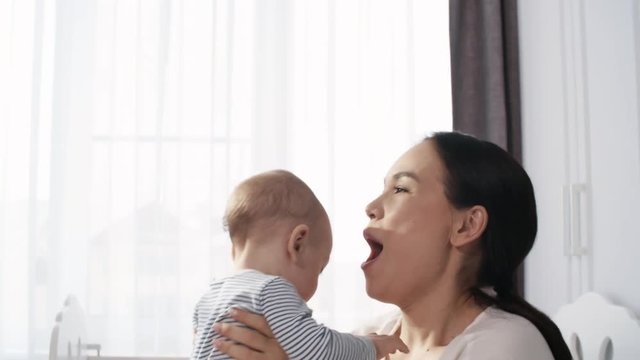 Chest-up shot of affectionate Asian mother playing with her baby, dressed in stripy bodysuit, lifting him up in her arms and rocking him, then gently kissing and cuddling
