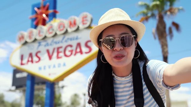 asian female travel blogger recording summer trip to america video by herself in selfie way. young girl doing coming gesture standing on sunny sky with welcome to fabulous las vegas sign background