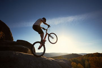 Naklejka na ściany i meble Silhouette of professional cyclist balancing on back wheel on trial bicycle, sportsman making acrobatic trick on the edge of rocky mountain. Blue sky and sunset on background. Concept of extreme sport