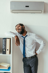 Fototapeta premium exhausted african american businessman standing under conditioner and holding hand on hip
