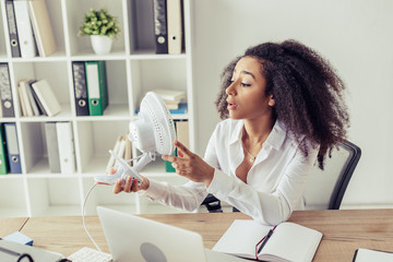 young african american woman holding desk fan while sitting at workplace and suffering from heat in office