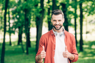 young man in red shirt standing in park with headphones on neck and showing thumbs up