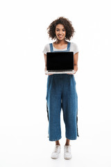 Image of brunette african american woman dressed in denim overalls smiling and holding open laptop