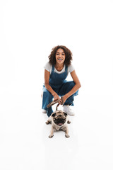 Image of delighted african american woman dressed in denim overalls squatting and poising with her pug dog