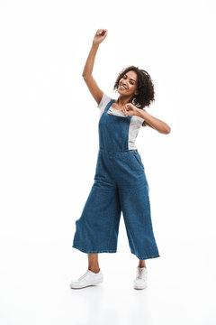 Image of joyful african american woman dressed in denim overalls smiling and dancing