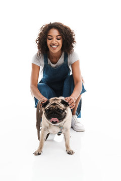 Image of happy african american woman dressed in denim overalls squatting and poising with her pug dog