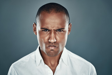 Obraz premium Feeling furious. Displeased young African man in shirt looking at camera and frowning while standing against grey background