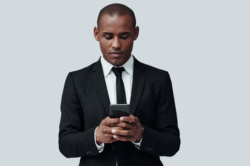 Checking income messages. Charming young African man in formalwear using smart phone while standing against grey background