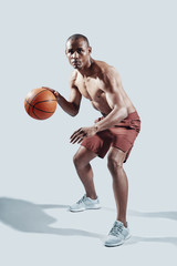 Naklejka premium Making effort. Full length of handsome young African man in sports clothing looking at camera while playing basketball against grey background