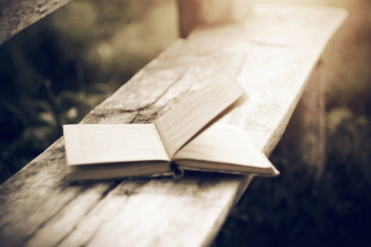 An old open paper book, with the wind turning the pages, lies on a wooden bench cracked by time.