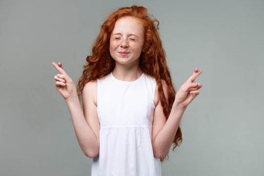 Portrait of dreamily nice little girl with ginger hair and freckles, stands with closed eyes and , crossed fingers and hopes for good luck, stands over gray background.