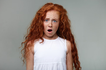 Close up of surprised girl with ginger hair and freckles, shocked looks at the camera wit wide open eyes and mouth, stands over gray background.