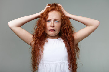 Naklejka premium Portrait of scared girl with ginger hair and freckles, holds his head with arms, looks at the camera wit digusted expression, looks shocked, stands over gray background.