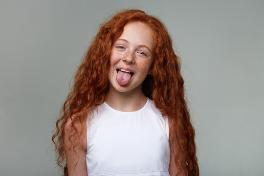 Photo of funny little girl with ginger hair and freckles, shows tongue at the camera, make grimace and stands over gray background, looks cute.