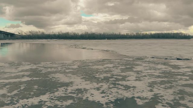 pictorial frozen river with patch of open water in ice against bank with dense forest bridge under cloudy sky upper view