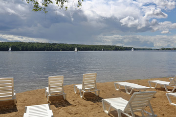 Empty plastic loungers on a sandy beach on the shore of a lake with sailboat