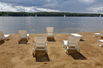 Empty plastic loungers on a sandy beach on the shore of a lake with sailboat