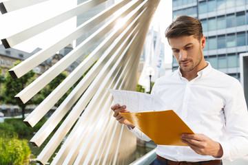 Concentrated handsome young business man standing near business center holding clipboard with...