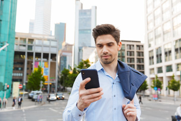 Young concentrated unshaved businessman walking outdoors on the street using mobile phone.