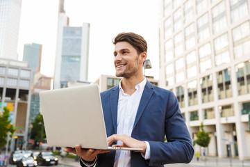 Cheerful happy handsome young business man standing near business center using laptop computer.