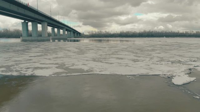 half frozen river water reflects grey bridge and clouds on patch of open water in ice on nasty winter day upper view