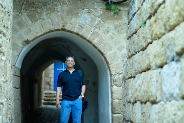 Fototapeta na wymiar Smiling trendy tourist senior with blue jeans and sunglasses standing by old ancient city street with brick historic stone wall.