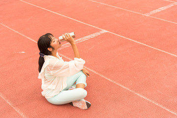 woman athlete sitting on runing track and drinking water from bottle. Cute slim young adult asian girl hold in hand bottle. Beautiful Female sitting on Red treadmill at the stadium and drinking water.