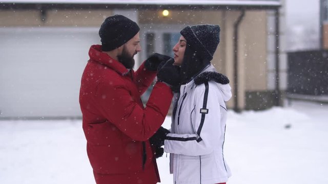 Bearded man and pretty woman kissing in winter backyard. Man trying to touch lips of girl with finger in glove, both laughing. Happy couple have a fun. Concept of outdoor recreation
