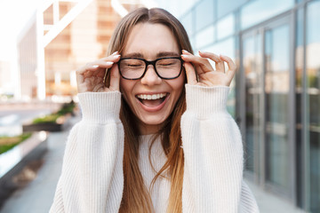 Beautiful young happy excited business woman posing walking outdoors near business center wearing eyeglasses.