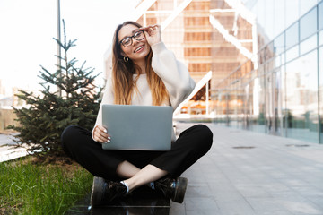 Beautiful young happy business woman posing sitting outdoors near business center wearing eyeglasses using laptop computer.