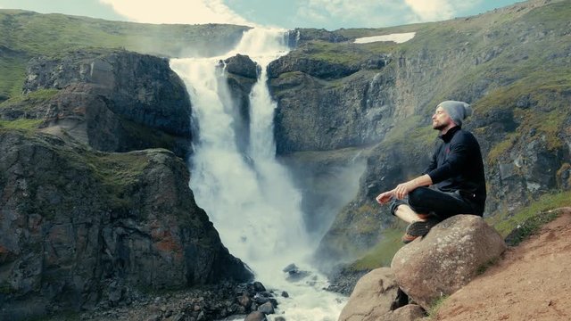 Man meditating on the rocks in Iceland. His arms are apart. He is sitting. Huge waterfall in the background.