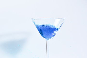 Cocktail with blue liquid in glass. Glass with blue water pouring with liquid with splashes and drops. Martini glass filling with alcohol with splashes on white background. Refreshing drink concept.