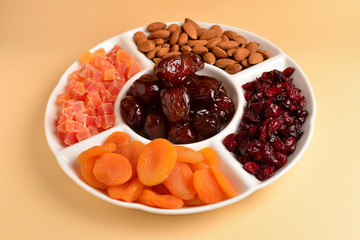 Mix of dried fruits and nuts on a white plate. Apricot, almond, raisin, dates fruit. On a brown background. Space for text or design.