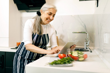 Smiling senior woman with white hair in in an apron using tablet while cooking at the kitchen