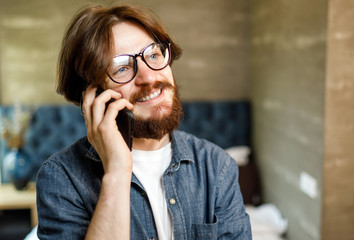Happy bearded hipster man wearing glasses speaking on the smartphone at the bedroom