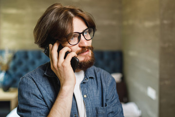Bearded hipster man wearing glasses speaking on the smartphone at the bedroom