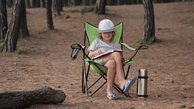 Child reading a book on pine wood background. Boy relaxing on chair in forest camp in summer vacation. Outdoors camping in childhood. Fresh air mood, beauty of nature concept. Open space