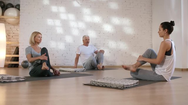 Full shot of middle-aged Caucasian husband and wife sitting together with crossed legs on mats in yoga studio, then teacher instructing them to bring one leg over other knee for ardha matsyendrasana