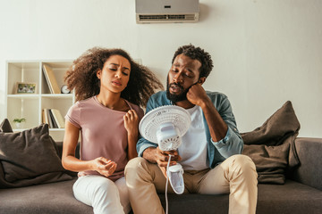 Fototapeta Unhappy African American man and woman holding blowing electric conditioner while suffering from summer heat at home obraz