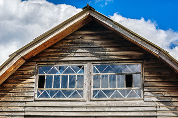 Gable End of an Aged Cottage with Horizontal Wooden Siding and Windows on a Sunny Day. White Clouds...