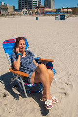 Mature baby boomer woman sitting on a colorful beach chair on a deserted beach on a sunny day. She has her hand on her head and appears to be deep in thought