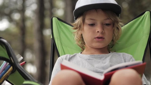 Portrait of child reading a book on pine wood background. Boy relaxing on chair in forest camp in summer vacation. Outdoors camping in childhood. Fresh air mood, beauty of nature concept. Open space