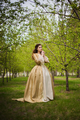 Beautiful girl in a gold dress against a background of green trees. A woman in a ball gown of the...