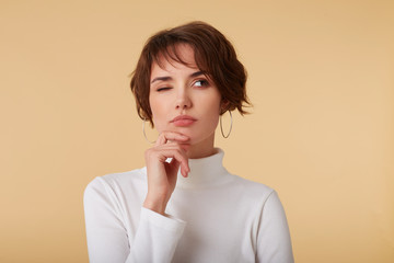 Close up of thinking short haired young woman wears in white golf, stands over beige background,...
