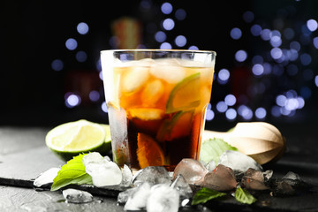 Glass with cola and lime slices, juicer, ice, mint against blurred lights background