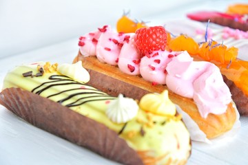 Tasty French eclairs with icing, cream, fresh berries and sugar decor elements. Set of delicious eclairs with creative colorful decor on white plates on neutral textured table. Homemade profiteroles 