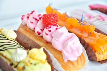 Tasty French eclairs with icing, cream, fresh berries and sugar decor elements. Set of delicious eclairs with creative colorful decor on white plates on neutral textured table. Homemade profiteroles 