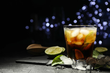 Glass with cola and lime slices, juicer, ice, mint against blurred lights background